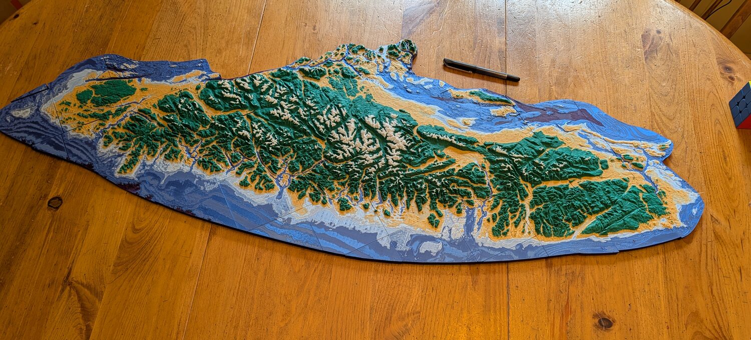 A one-metre-long 3D-printed relief map of Vancouver Island assembled on a wooden table, coloured in bands from deep purple ocean through blues, gold lowlands and green mountains to white peaks. A pen lies beside it for scale.