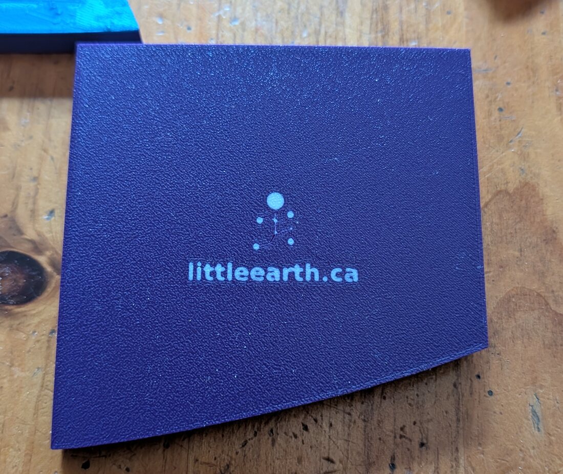 The underside of a printed tile: a flat dark base with the Little Earth logo and the words littleearth.ca picked out in white, and one curved edge where the tile meets the boundary.