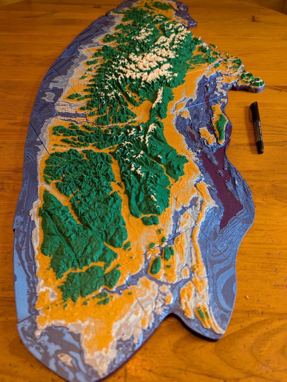 An oblique view along the printed island, showing green mountains and gold lowlands on the left and, on the right, the Strait of Georgia rendered as a deep purple trough flanked by pale blue shelves.