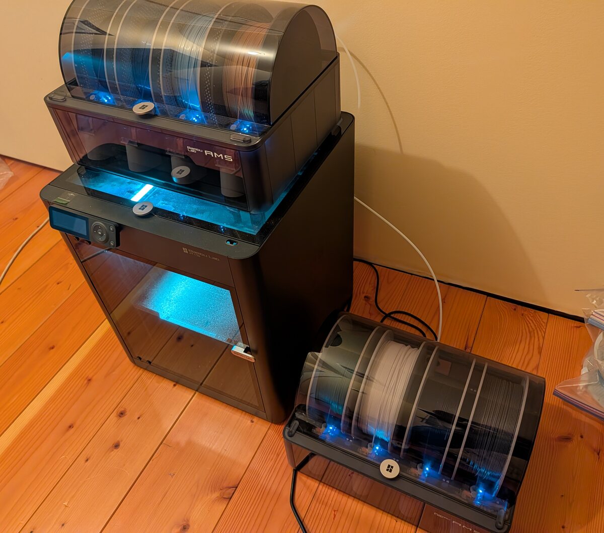 A Bambu Lab P1S 3D printer glowing blue, with one four-spool AMS filament changer stacked on top of it and a second AMS unit sitting on the floor alongside, loaded with white and dark filament.