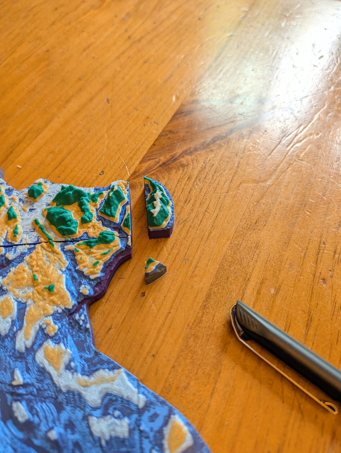 Two very small printed fragments sitting on a wooden table beside the edge of the main model — one a narrow crescent, the other a chip barely a centimetre across.